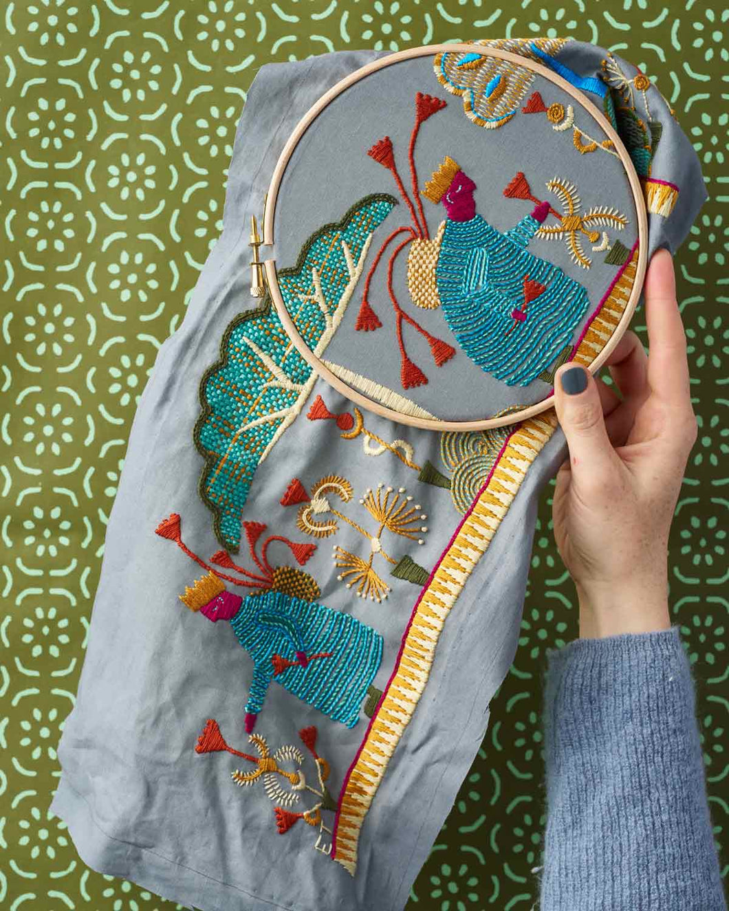 Embroidery hoop with colorful floral design held over a piece of embroidered fabric on a patterned background.
